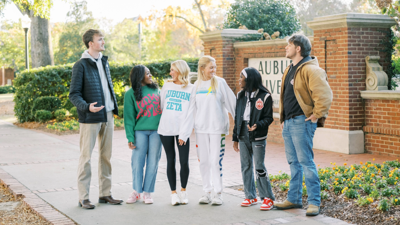 Six students stand chatting on a campus sidewalk in front of a brick sign that reads “Auburn University,” with trees, greenery, and flowers around them.