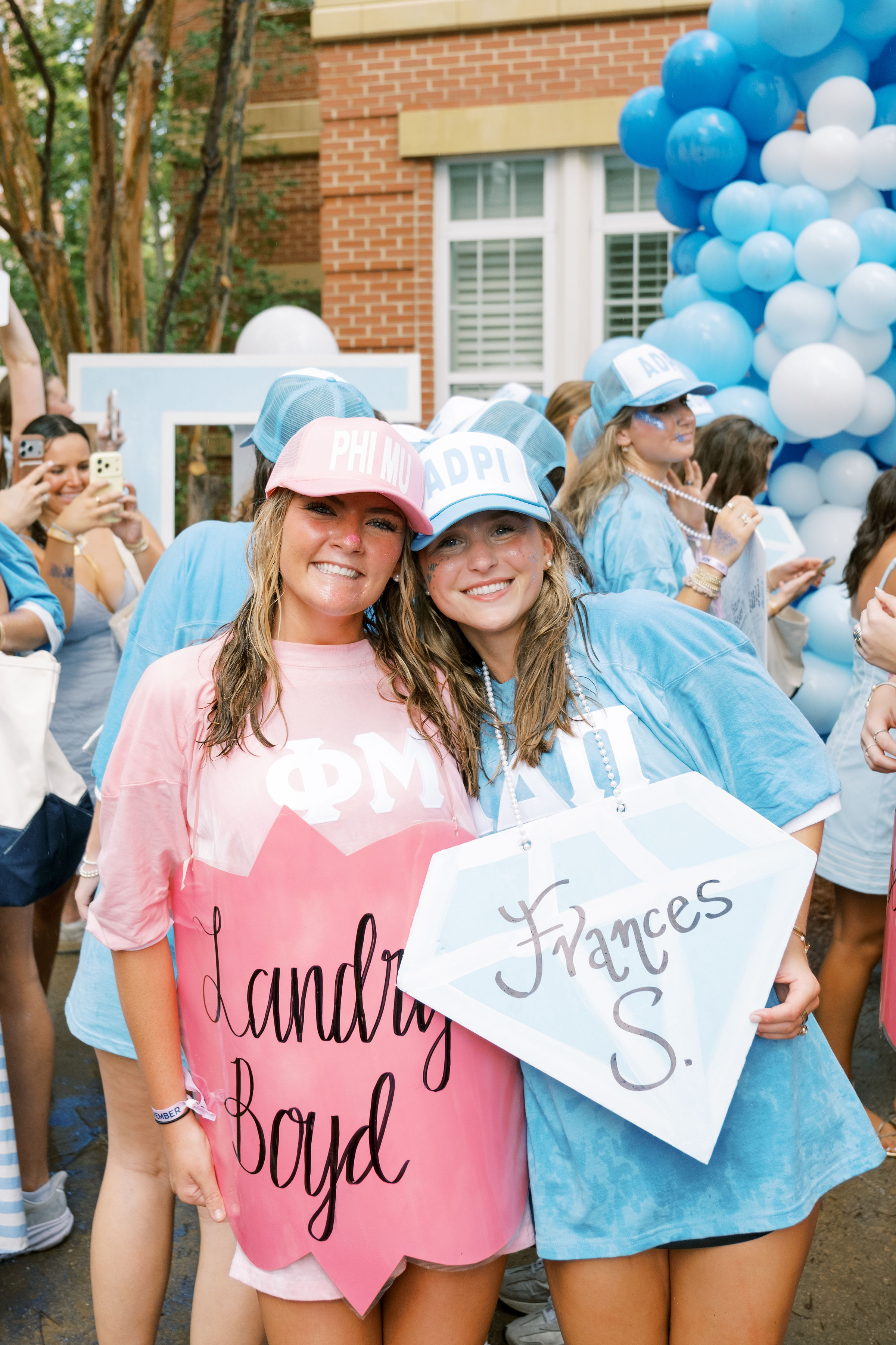 Two students wearing sorority-themed shirts and caps pose together at an outdoor bid day celebration, surrounded by other participants and blue-and-white balloon decorations.