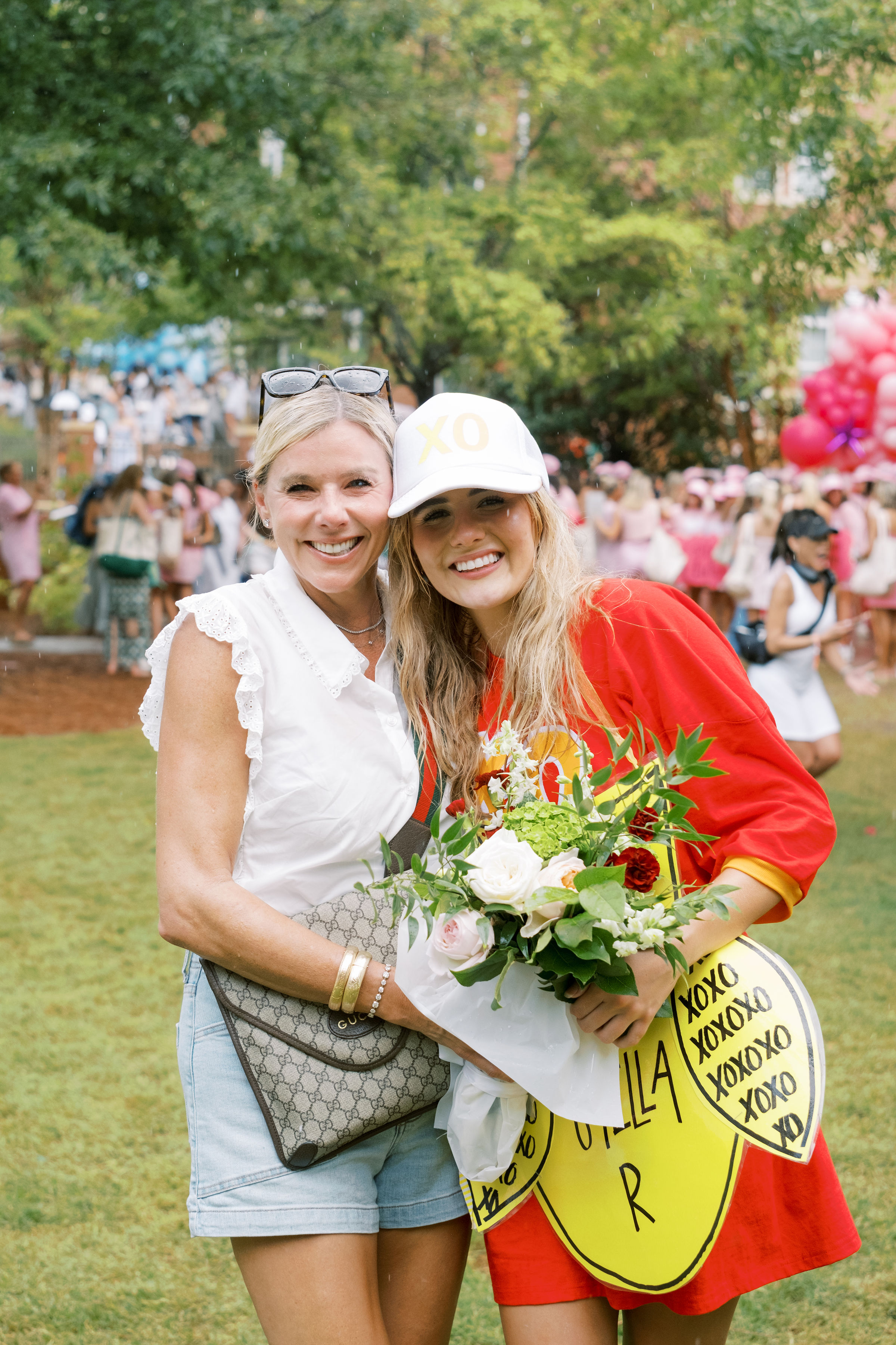 Two people pose together outdoors during a sorority bid day celebration, holding a bouquet of flowers. One person wears a white XO cap and a red shirt with a large decorative name tag, while a crowd and pink balloons fill the background.