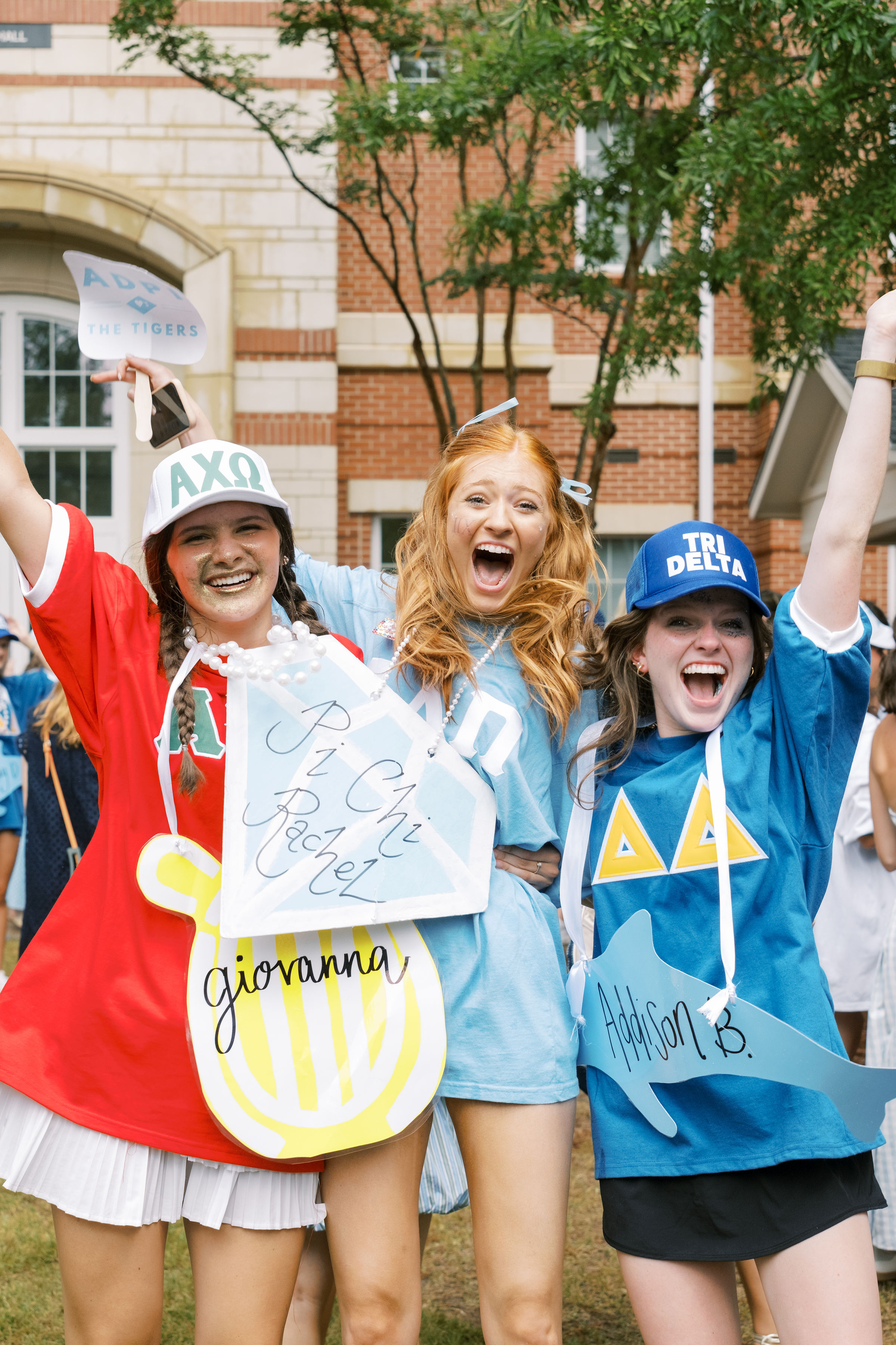 Three students in sorority-themed shirts and hats pose with raised arms and handmade signs during an outdoor campus recruitment celebration.