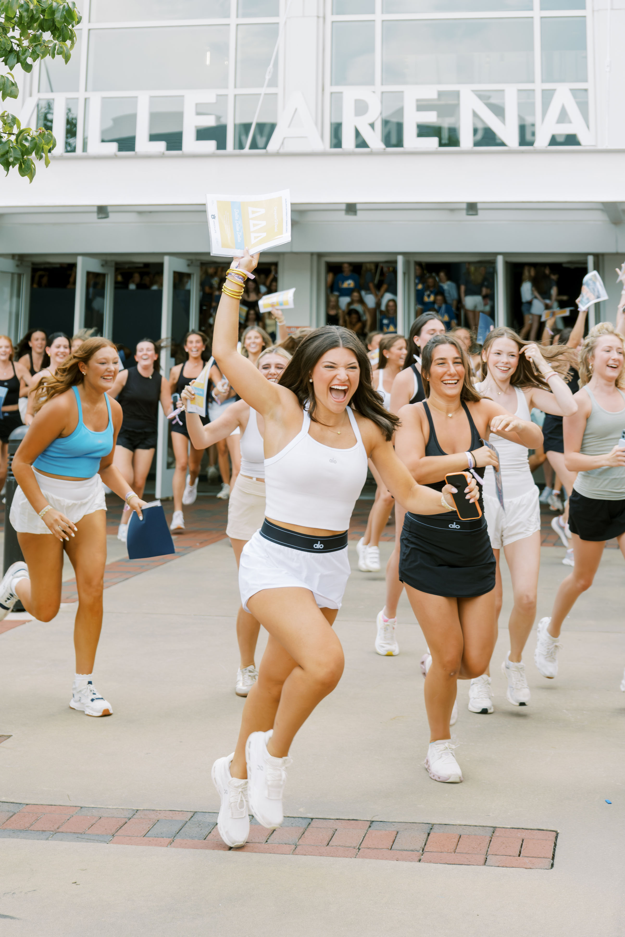 Students run from a campus building during bid day, waving papers and gathering outside for a sorority recruitment celebration.