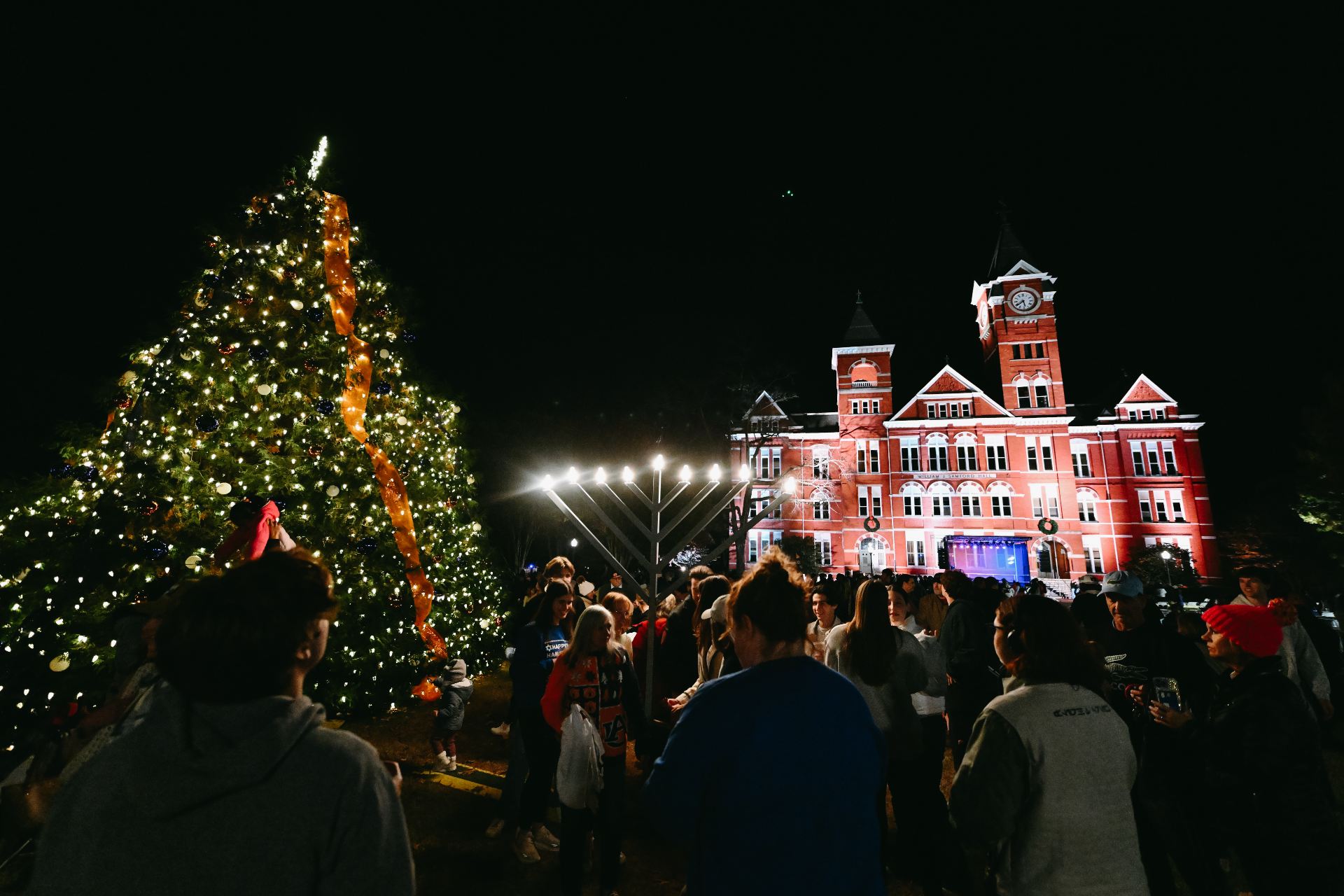 students and community members in front of a large tree with lights and a Menora in front of Samford Hall at night