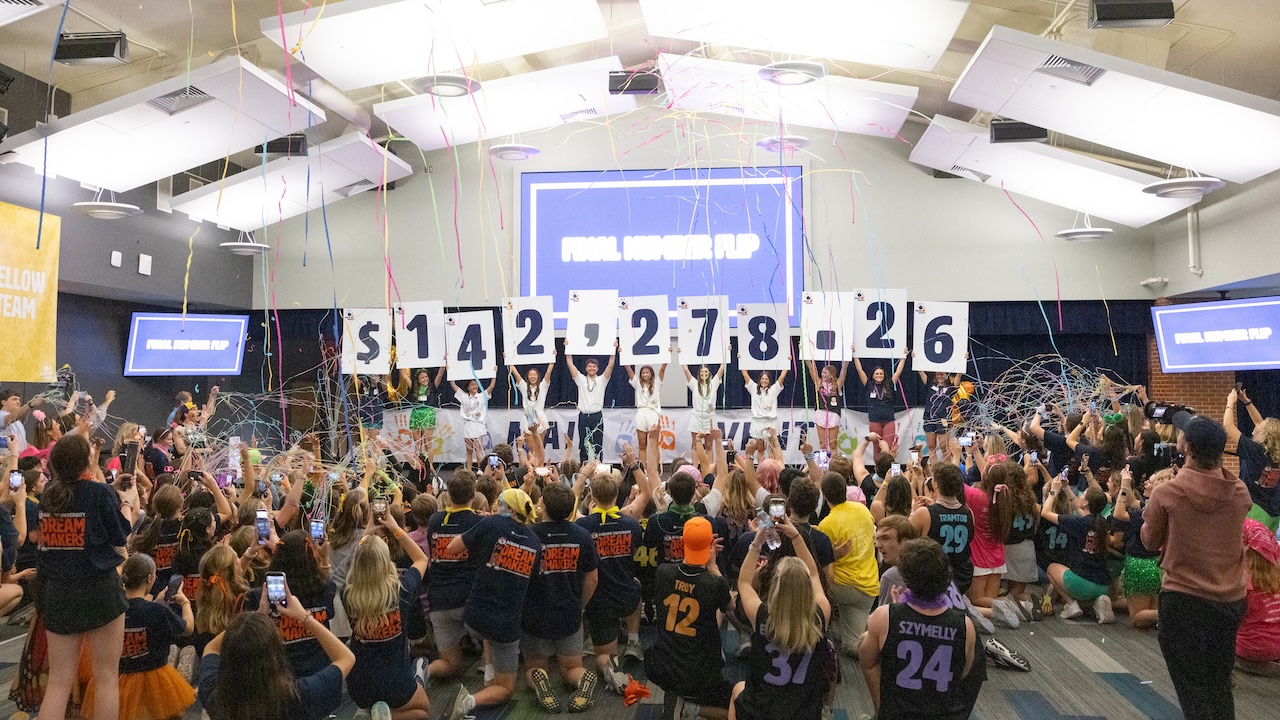 Students in AU Dream Makers on stage in a large room surrounded by other students holding signs on a stage that say $142,278.26 
