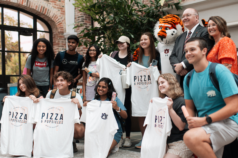 Students posing with President Roberts and Aubie while holding tshirts that say Pizza and Popsicles with the president